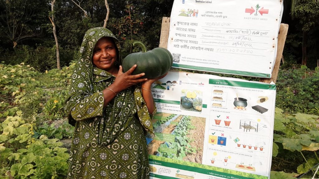 A woman farmer in Bangladesh holds a pumpkin by the signboard for her demo plot