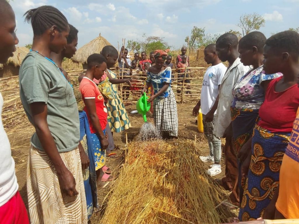 A woman demonstrates to assembled trainees how to water a ground nursery for onions