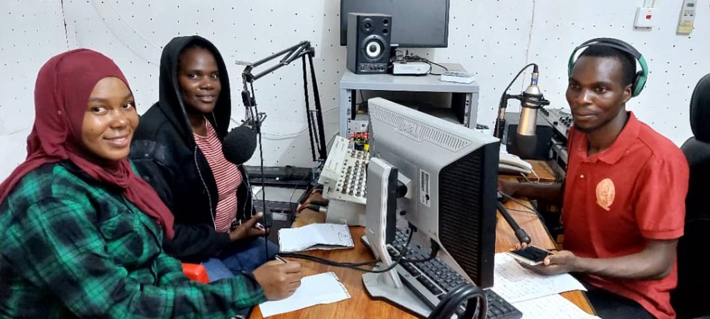 EWS-KT Technical Field Officers Hadija Lihumbo and Lydia Mkopa and radio presenter Mwl. Natabu sit at a table with microphones and other radio equipment.