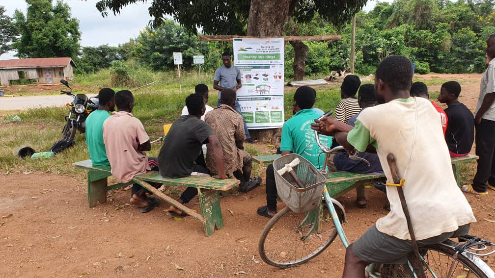 A man on a bicycle watches a group of farmers listening to EWS-KT Technical Field Officer Mark Nabre.