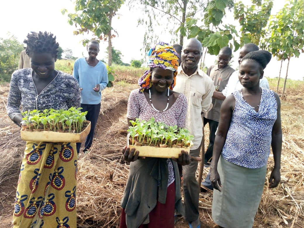 Two women carry trays of seedlings growing in leaf pots as fellow farmer look on.
