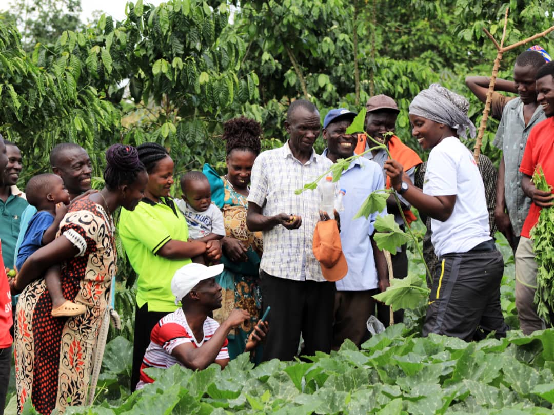 Female Technical Field Officer holds a pumpkin vine with large group of female and male farmers looking on.