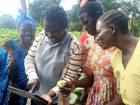 Female Technical Field Officer sharing technological resources on a tablet, surrounded by women farmers.