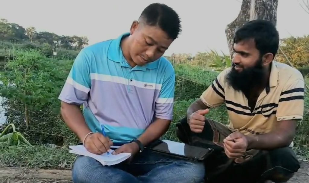 Technical Field Officer Miltan Chakma and farmer Md. Oliullah sit on the ground while discussing farm costs and updating crop data in EWS-KT’s data-tracking app. Miltan writes on some paper on one leg, while a tablet rests on his other leg.