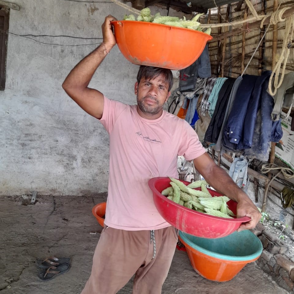 Farmer Sanjay Patidar holds a large container of bitter gourd fruit in one hand and another container on his head.