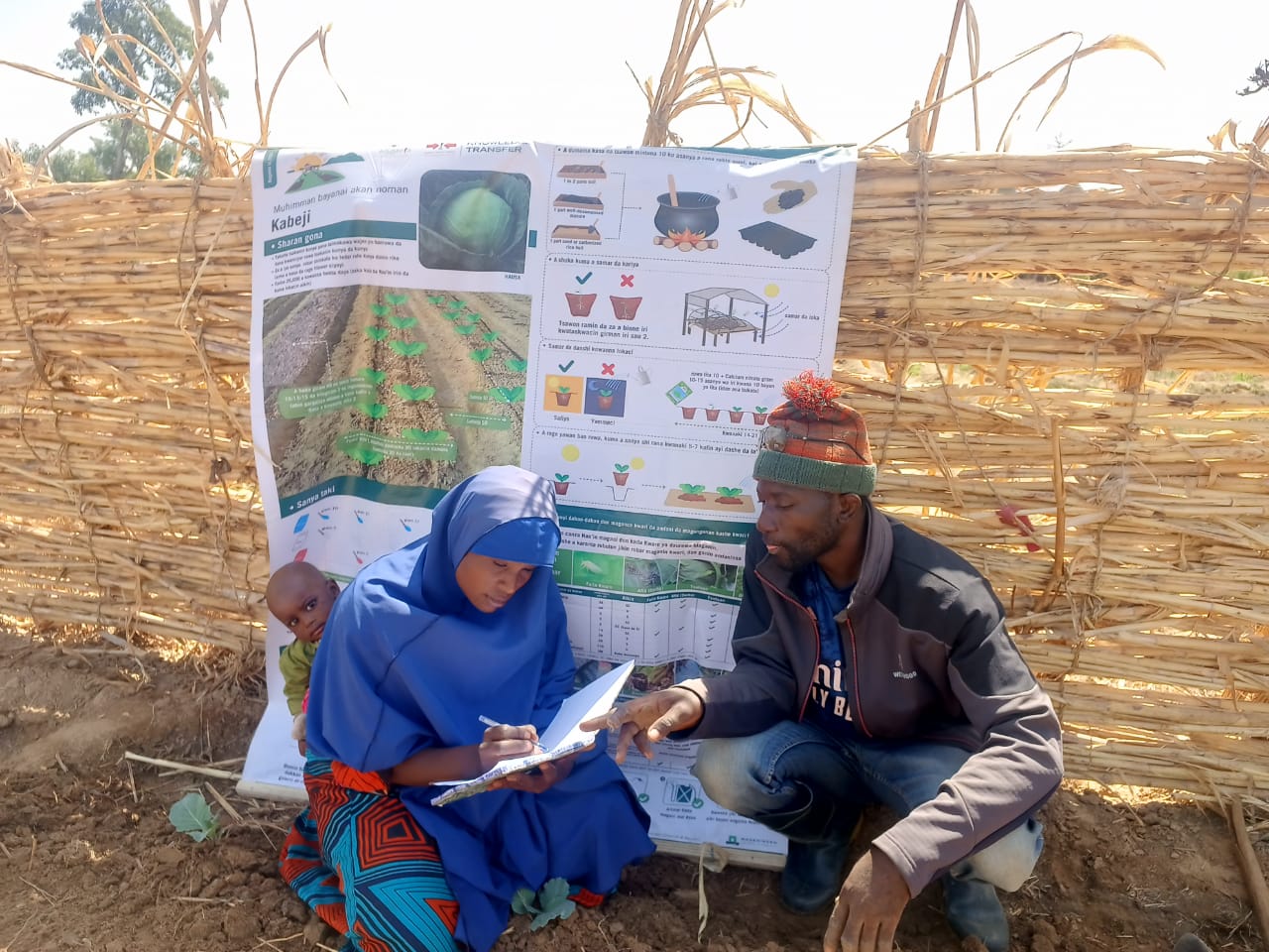 Male EWS-KT Technical Field Officer assists a female key farmer with farm record-keeping. Behind them is an informational banner.