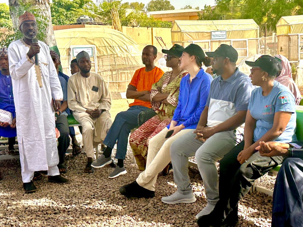 Dr. Miswaru Bello, Deputy Vice Chancellor of Sa’adatu Rimi University of Education, stands as he addresses visitors to the learning site at the university. Seated on a bench facing him are Mohammed Salasi Idris, HortiNigeria Program Director;  Temitayo Akinbiyi, Policy Officer, Education, and Mariska Lammers, First Secretary, Food Security and Climate, from the Embassy of the Kingdom of the Netherlands in Abuja; Yusuf Dramani, Country Director for IFDC Nigeria; and Ruth Ardzard, EWS-KT Knowledge Transfer Manager for Nigeria.
