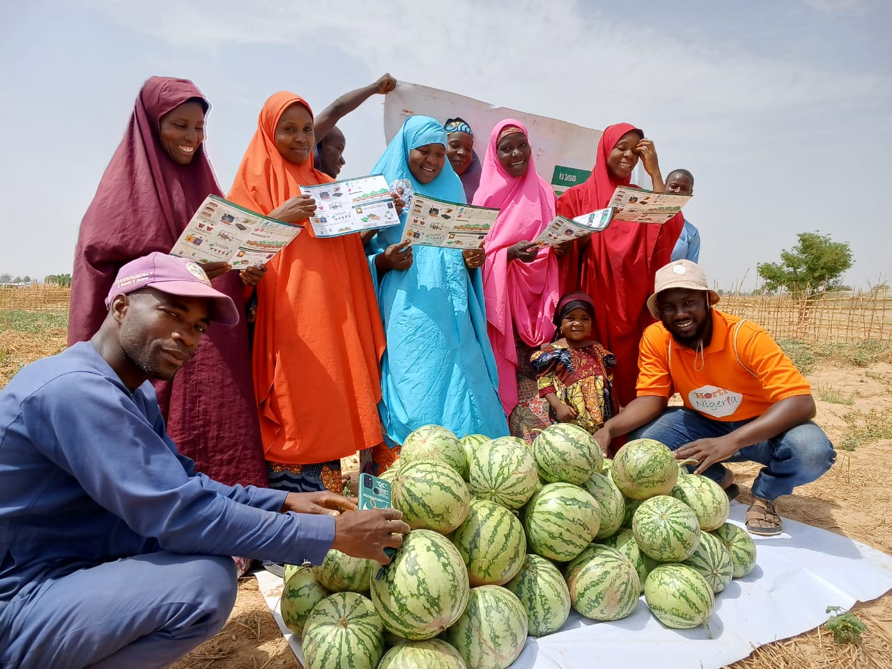 Women holding crop guides stand in front of a pile of watermelons, with men flanking the watermelons.