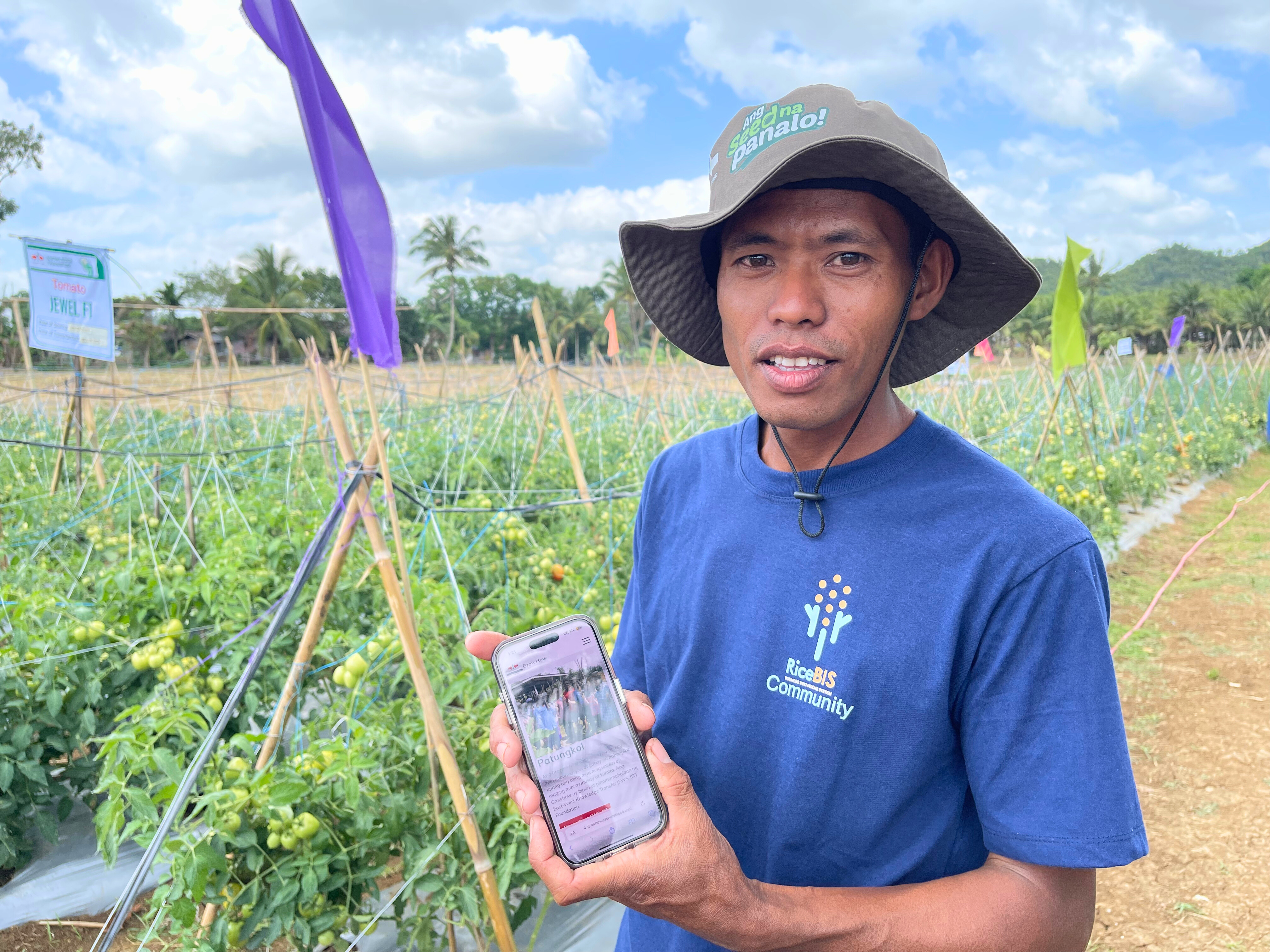Young farmer Ronel Cañetan Lausa stands in front of his tomato field. He is holding the mobile phone through which he first connected with EWS-KT.