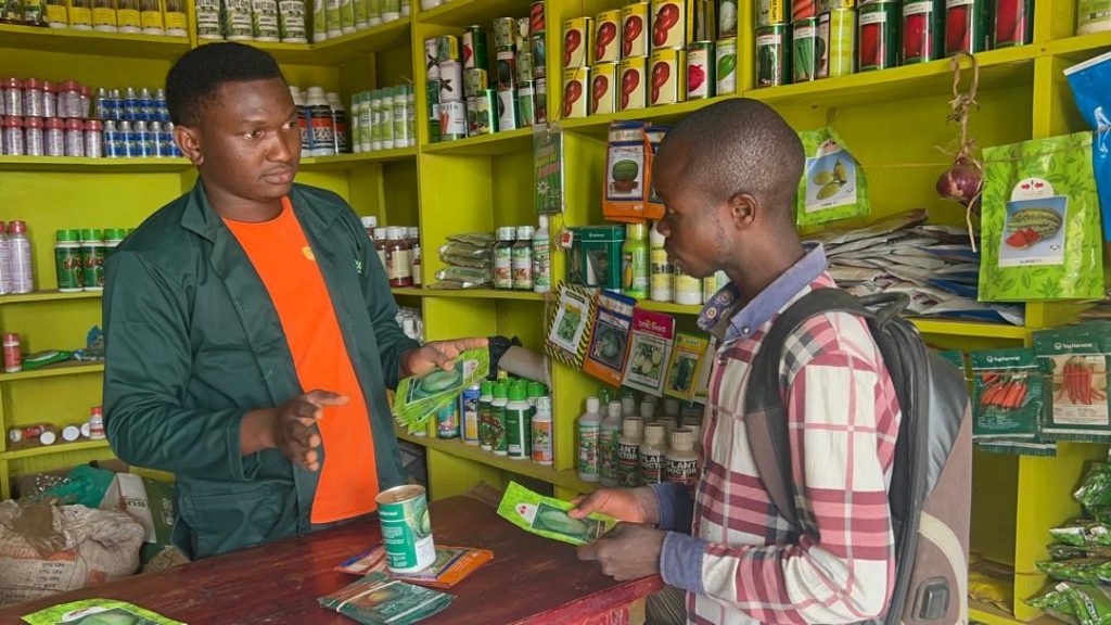 Standing behind the counter in his agro-input shop, Musasizi Moses talks to a customer.