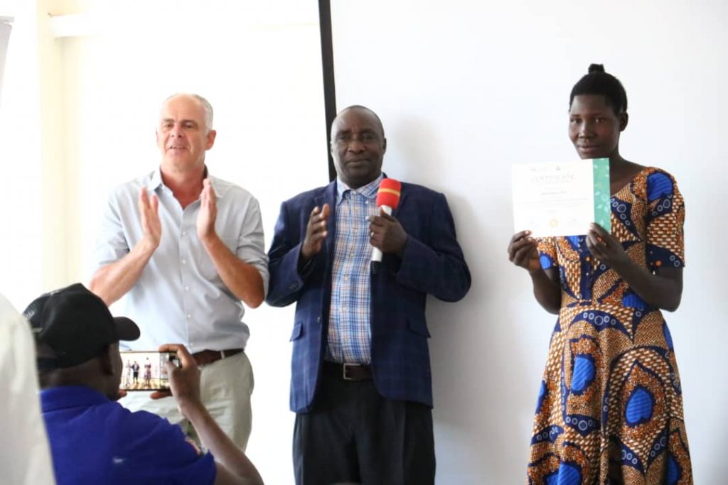 EWS-KT Executive Director Stuart Morris and Lira District Production and Marketing Officer Thomas Okello clap as a female farmer holds up her certificate.