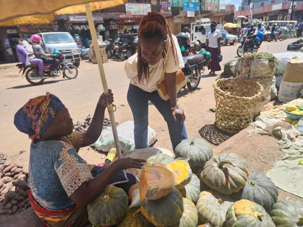 EWS-KT Technical Field Officer Christine Ategeka talks with market seller Ssenga, who is displaying pumpkins in the market beneath an umbrella.