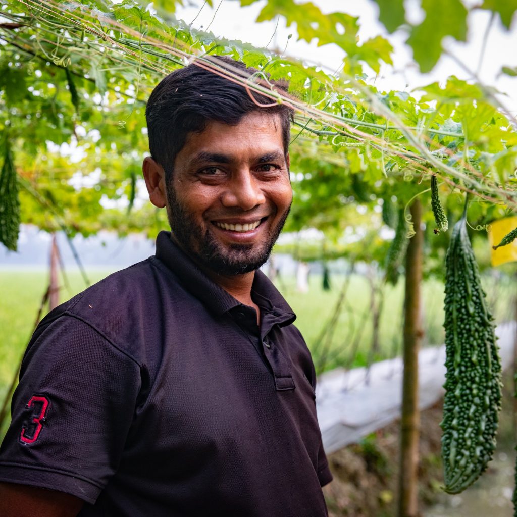 Farmer Md. Jahangir smiles as he stands next to a row of bitter gourd.
