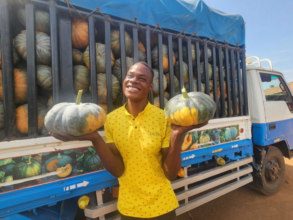 Ugandan farmer Robert Asiimwe smiles broadly as he holds a pumpkin in each hand in front of truck filled with pumpkins.