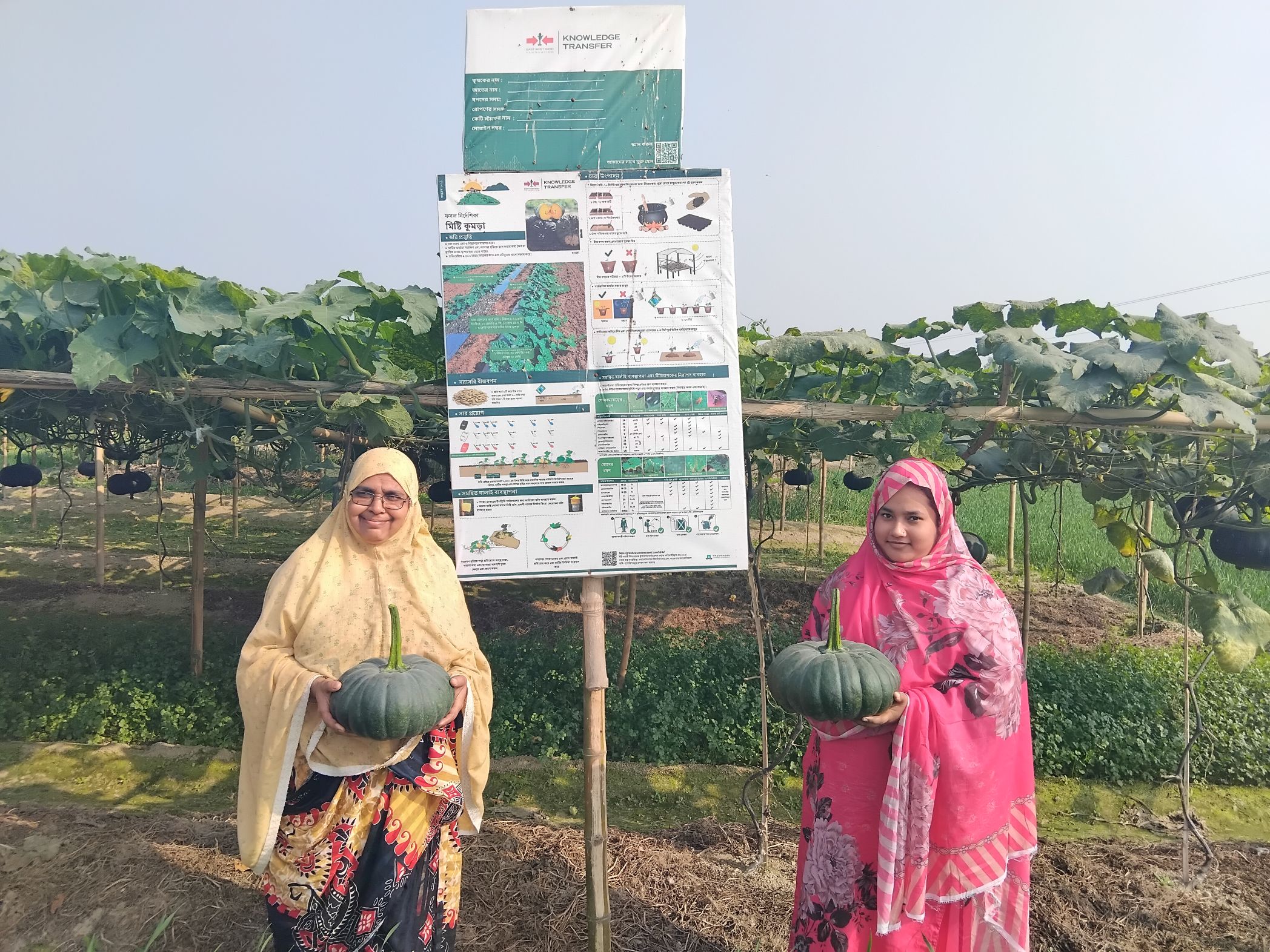 Two female farmers hold pumpkins in front of a pumpkin demonstration field.