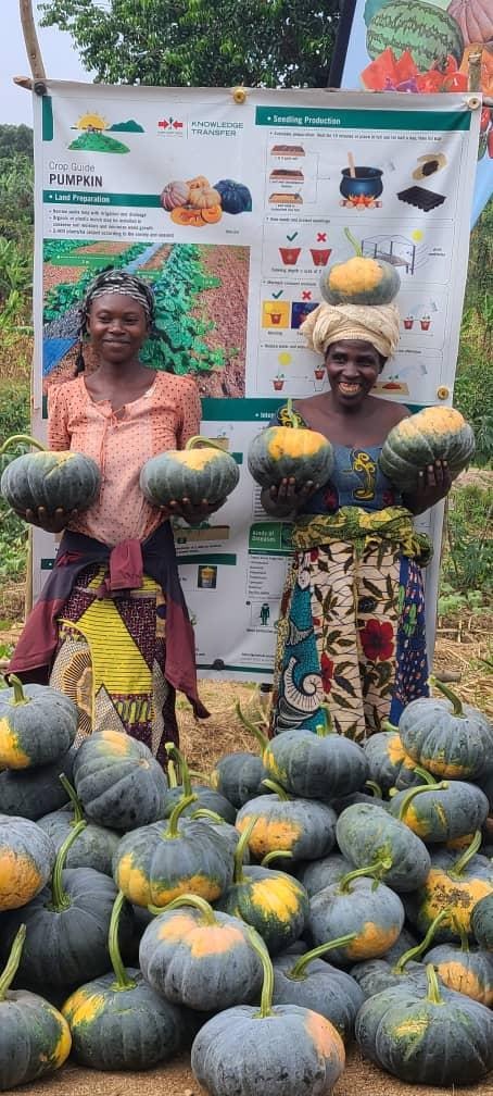 Ugandan pumpkin farmer Odeta Dukizmana poses with another women in front of an EWS-KT banner on pumpkin farming. In front of them are a pile of pumpkins.
